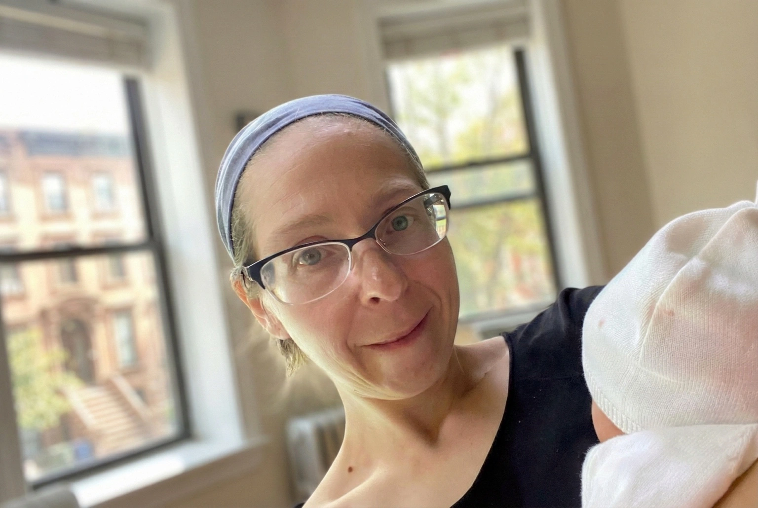 A postpartum doula with glasses smiles gently while holding a newborn in a sunlit Brooklyn apartment.