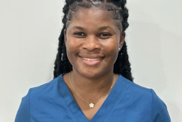 Sydia, postpartum doula with over 13 years of experience, smiling and wearing blue scrubs against a light background.