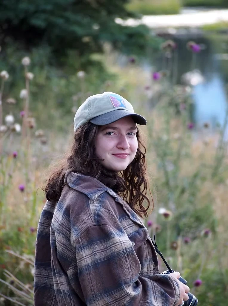 Portrait of Zoe Deenihan, a postpartum doula and lactation educator, smiling outdoors in a natural field setting while holding a camera.